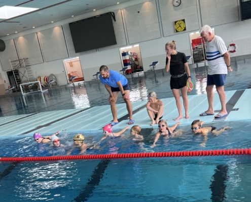 Kinder im Schwimmbecken und drei Trainer an Land
